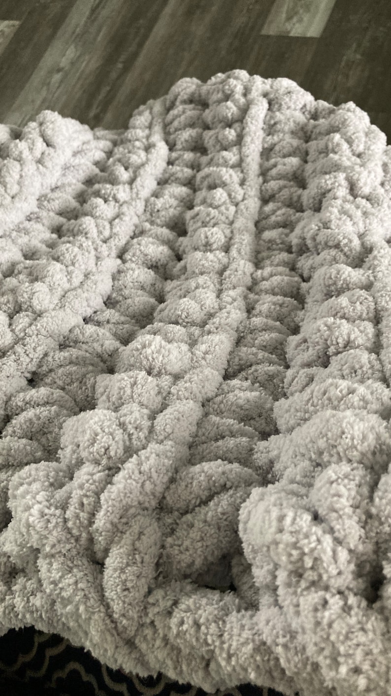 Peut inclure: Une couverture en tricot moelleuse, &eacute;paisse et grise avec un motif tiss&eacute;. La couverture est faite de fil &eacute;pais et a une texture duveteuse.
