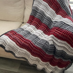 May include: A crocheted blanket featuring a repeating striped pattern in shades of deep red, charcoal gray, light gray, and cream. The textile has a textured, ribbed stitch construction with a wavy, scalloped edge along the bottom border.