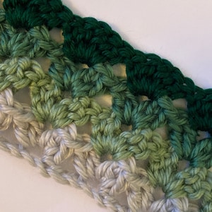 May include: A crocheted fabric strip with a gradient of green and white yarn. The fabric is made with a repeating pattern of squares.
