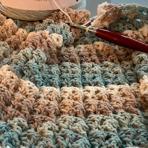 May include: A close-up of a crocheted blanket in shades of blue, brown, and white. The blanket is made with Caron Cotton Cakes yarn. A crochet hook is lying on top of the blanket.