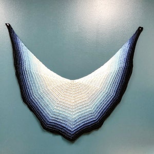 May include: A crocheted shawl with a gradient of blue, white and black colours. The shawl is shaped like a crescent moon and has a scalloped edge.