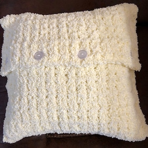May include: A white crocheted pillow with three buttons. The pillow is made with a fluffy yarn and has a textured surface.