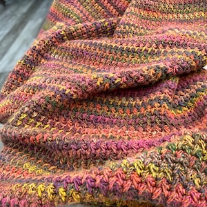 May include: Close-up of a hand-crocheted scarf in shades of pink, orange, yellow, and green. The scarf has a textured, woven appearance with a diagonal pattern. The soft, folded fabric is visible, showcasing the intricate stitch work.
