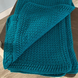 May include: A teal blue crocheted blanket folded on a wooden chair. The blanket has a textured, woven pattern.