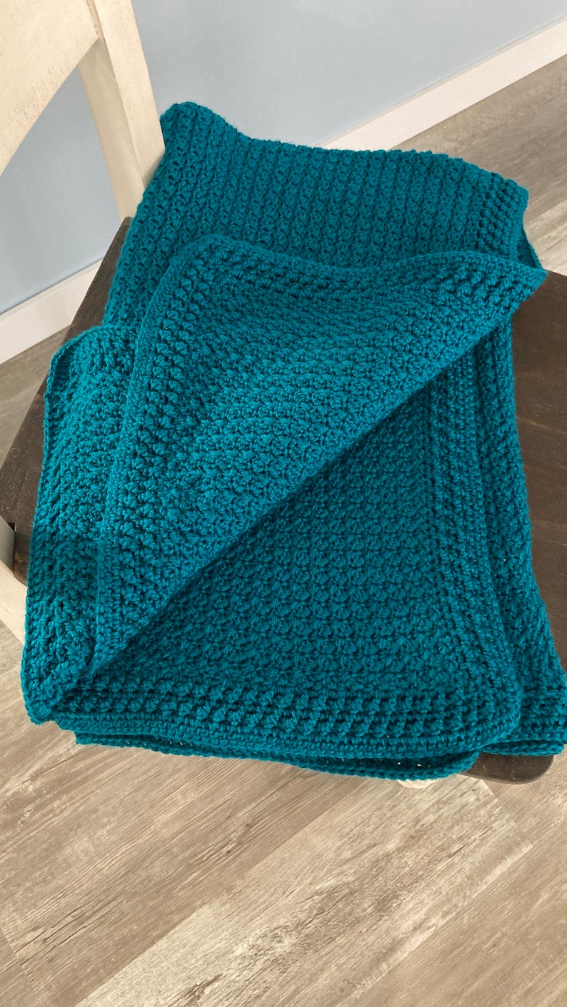May include: A teal blue crocheted blanket folded on a wooden chair. The blanket has a textured, woven pattern.