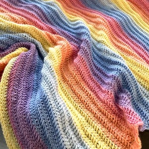 Op de afbeelding: Een handgemaakte, kleurrijke gehaakte deken. De deken heeft een levendige regenboog van kleuren, waaronder geel, oranje, roze, paars en blauw. Het getextureerde steekpatroon voegt visuele interesse toe. De deken is gevouwen en gedrapeerd, wat de zachte textuur laat zien.