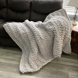 Peut inclure: Une couverture en tricot moelleuse, &eacute;paisse et de couleur gris clair, drap&eacute; sur un canap&eacute; brun. La couverture a un motif en tricot torsad&eacute; textur&eacute;.