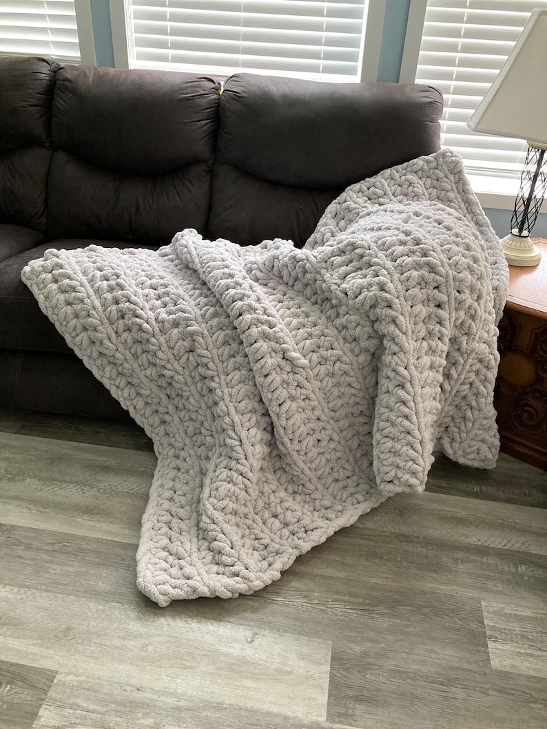Peut inclure: Une couverture en tricot moelleuse, &eacute;paisse et de couleur gris clair, drap&eacute; sur un canap&eacute; brun. La couverture a un motif en tricot torsad&eacute; textur&eacute;.