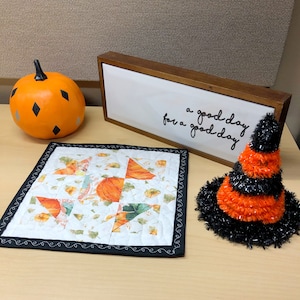 May include: A white quilted fabric square with a black border and a patchwork design of orange and green fall leaves. The fabric square is on a wooden table with a black and orange witch's hat and an orange pumpkin.