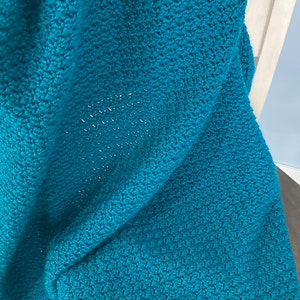 May include: A teal blue crocheted blanket with a textured pattern. The blanket is folded and draped over a wooden chair.