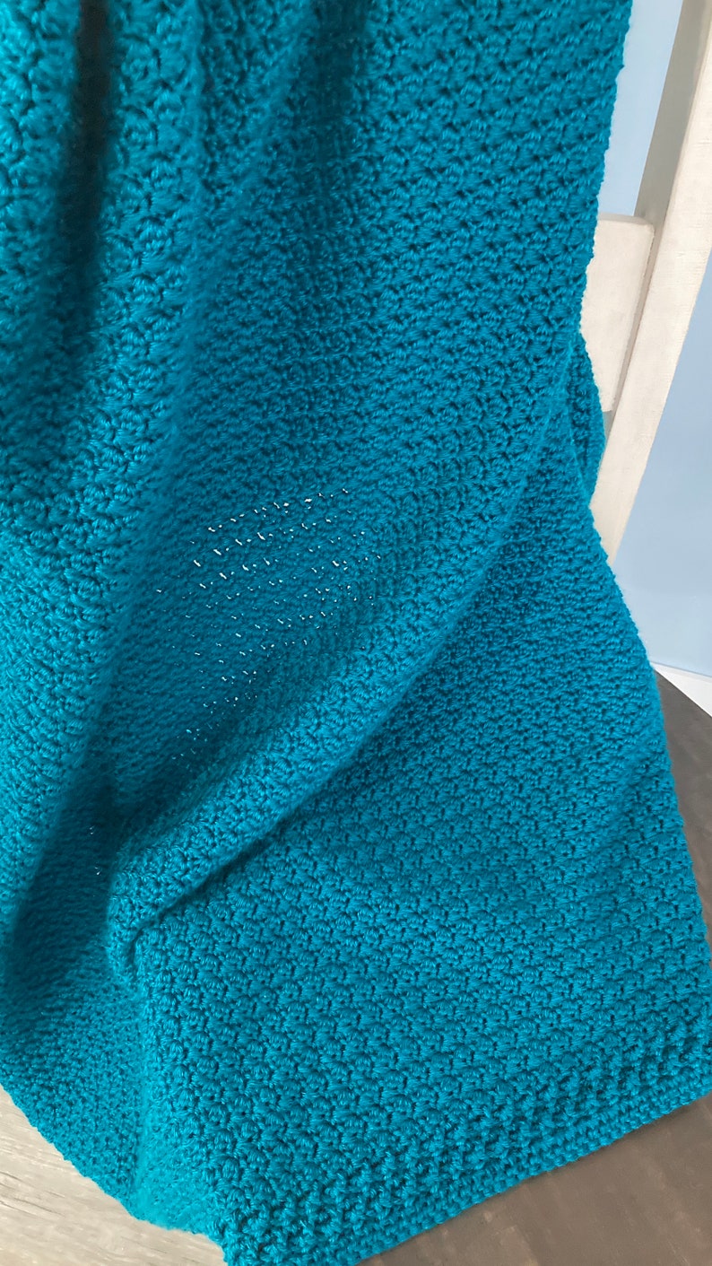 May include: A teal blue crocheted blanket with a textured pattern. The blanket is folded and draped over a wooden chair.