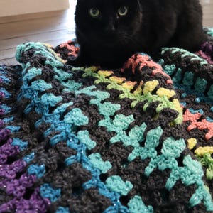May include: A black cat rests on a colorful, handmade crochet blanket. The blanket features a dark gray base with vibrant accents of purple, blue, turquoise, yellow, and pink. The crochet pattern is visible, adding texture.