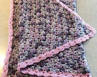 May include: A crocheted baby blanket in shades of purple, pink, and gray with a pink crocheted border.