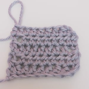 May include: Close-up of a crocheted swatch in shades of lavender and gray. The handmade fabric features a textured stitch pattern with visible loops and a loose end of yarn. The background is plain white.