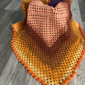 May include: A crocheted baby blanket with a heart design in shades of orange, yellow, and brown. The blanket has a crocheted border with orange pom-poms.