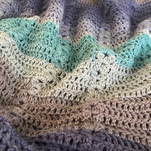 May include: A handmade crochet blanket in shades of blue, gray, and teal. The blanket features a textured stitch pattern, creating a soft and cozy appearance. The color palette includes various shades of blue, gray, and teal, creating a visually appealing design.