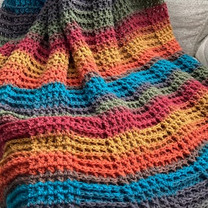 May include: A handmade crochet blanket featuring a textured waffle stitch pattern. The yarn displays a vibrant gradient of colors including deep charcoal, olive green, mustard yellow, bright orange, and turquoise blue arranged in horizontal stripes.