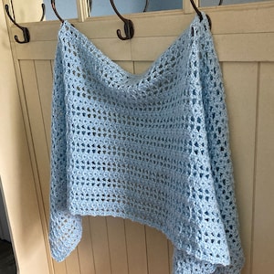 Puede incluir: Un chal de ganchillo azul claro colgado de un gancho. El chal tiene un delicado patrón de calado.