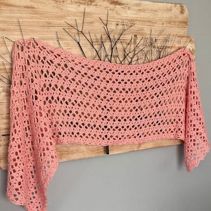 Peut inclure: Un châle en crochet rose avec un motif ajouré délicat. Le châle est drapé sur une surface en bois avec un motif métallique décoratif.