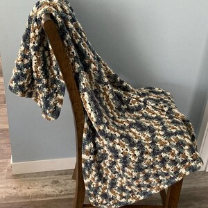May include: A crocheted blanket in blue, brown, and cream, draped over a wooden chair. The blanket's textured pattern is visible. The chair has a curved back and legs. The blanket is folded over the seat of the chair.