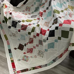 May include: A white quilt with a patchwork design featuring squares of various colors and patterns, including red, green, blue, and black. The quilt is folded and draped over a brown couch.