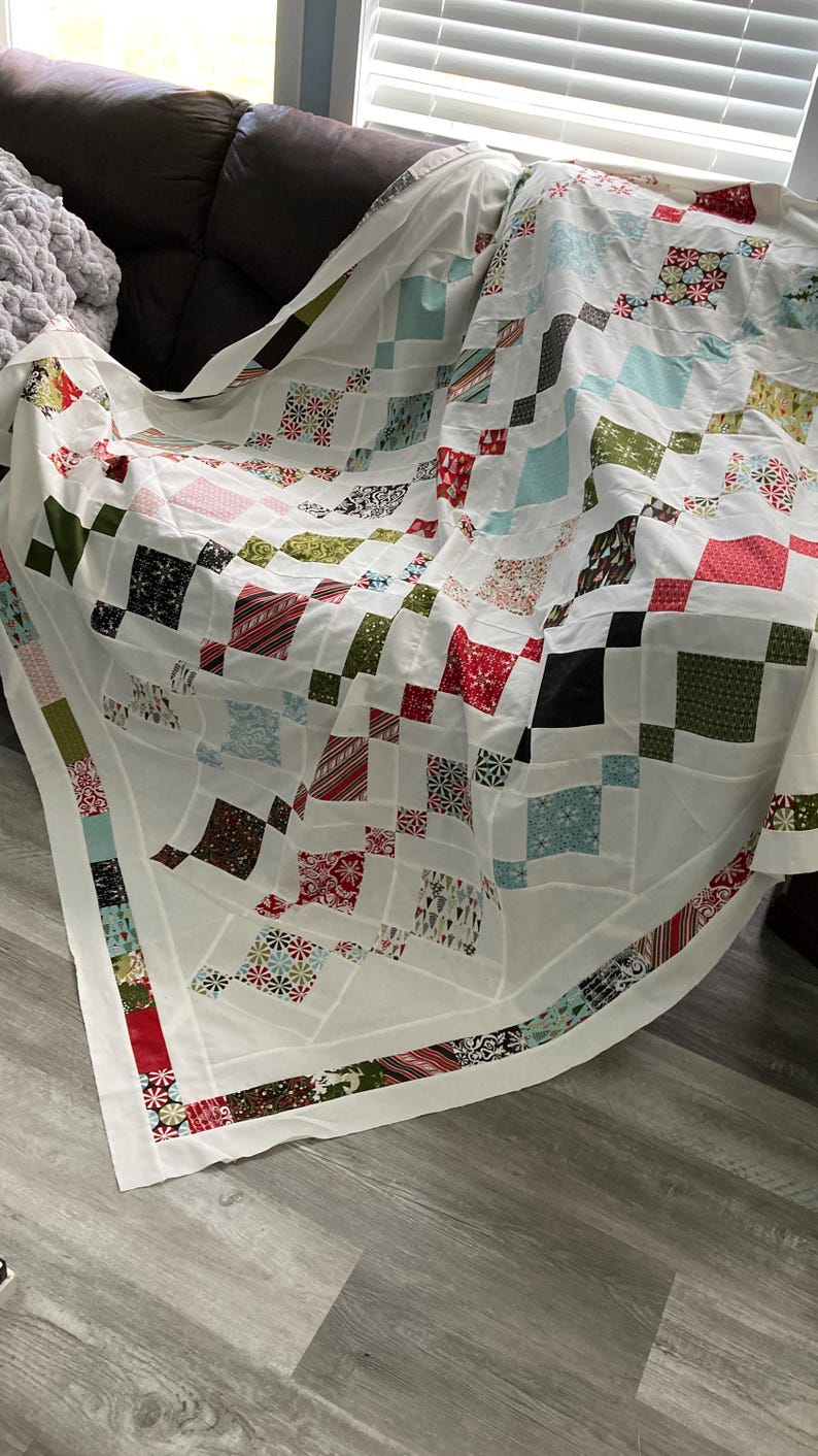 May include: A white quilt with a patchwork design featuring squares of various colors and patterns, including red, green, blue, and black. The quilt is folded and draped over a brown couch.