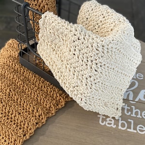 Puede incluir: Artículos de ganchillo hechos a mano en tonos beige y marrón. Un paño crema doblado y un mantel individual marrón se exhiben sobre una superficie de madera. Una cesta de metal oscuro contiene un paño marrón enrollado. Los artículos tienen una apariencia texturizada y tejida.