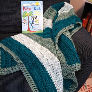 May include: A green, white, and gray striped crocheted blanket is draped over a black armchair. A children's book titled "Pete the Cat" is sitting on the blanket.