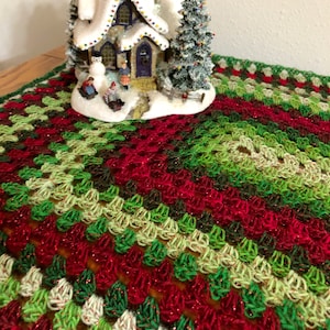 Puede incluir: Una manta de crochet roja, verde y blanca con una escena de pueblo de invierno nevado en el centro. El pueblo cuenta con una pequeña casa con una ventana iluminada, un muñeco de nieve y un árbol de Navidad.