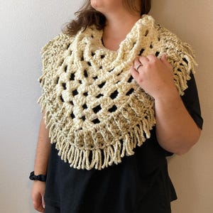 May include: A cream-colored crochet cowl with a chunky, open-weave diamond pattern and a thick fringe border. The yarn features subtle brown flecks throughout the texture, creating a variegated appearance across the triangular-shaped garment.
