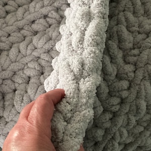 Peut inclure: Gros plan sur une couverture en tricot grise &agrave; la texture moelleuse. La couverture est pli&eacute;e en deux et une main tient le bord de la couverture.