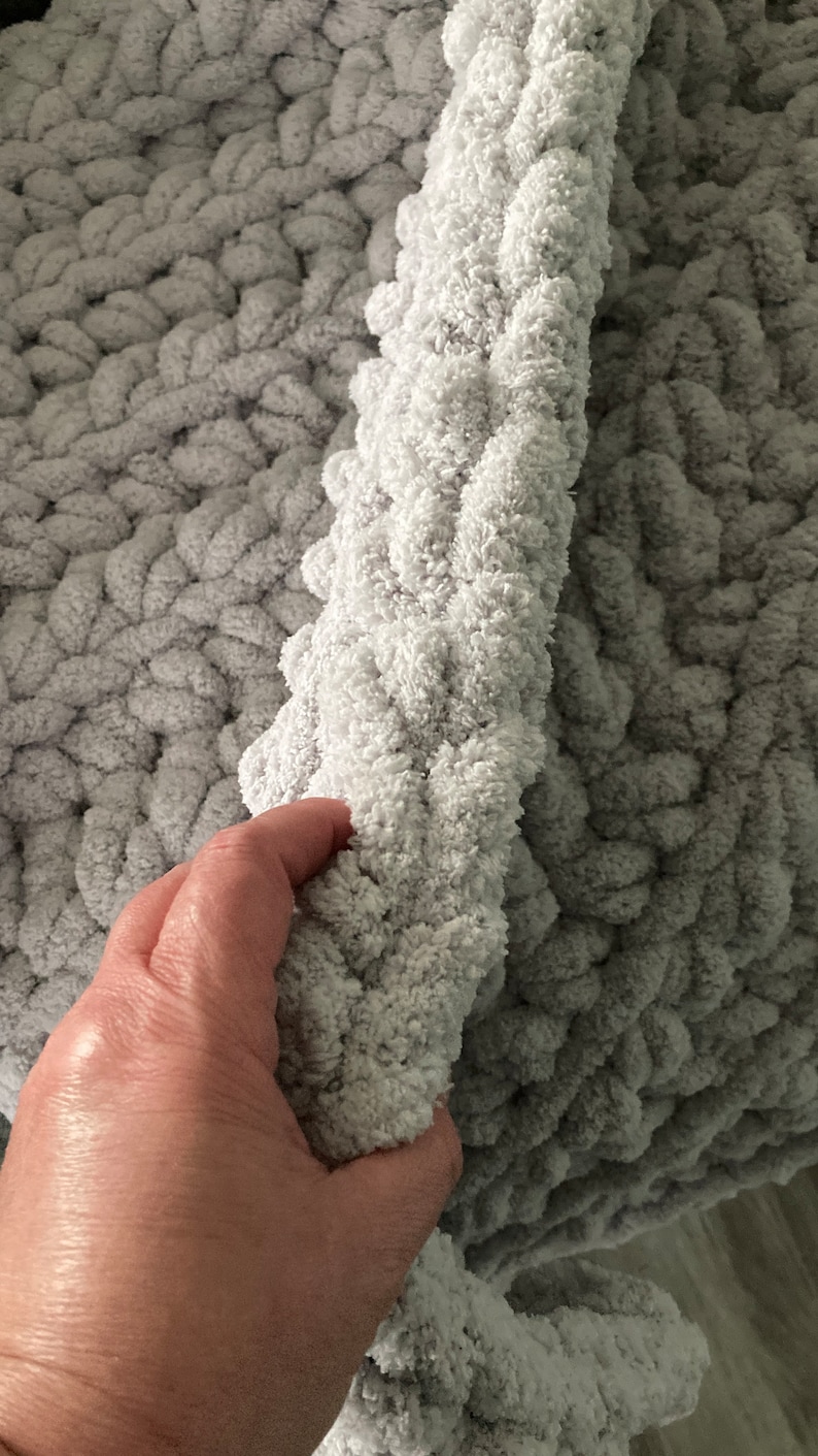 Peut inclure: Gros plan sur une couverture en tricot grise &agrave; la texture moelleuse. La couverture est pli&eacute;e en deux et une main tient le bord de la couverture.