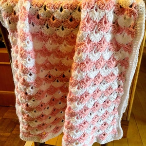 May include: A pink and white crocheted baby blanket with a scalloped edge. The blanket is draped over a wooden chair.