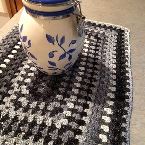 May include: A white ceramic cookie jar with a blue lid and blue floral design. The jar is sitting on a black and white crocheted placemat.
