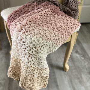 May include: A hand-crocheted blanket in a gradient of colours, from light beige to white, pink, and brown. The blanket is draped over a small, cream-coloured chair with curved legs, showcasing its textured pattern.