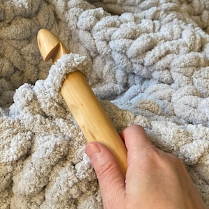 Peut inclure: Gros plan sur un crochet en bois utilis&eacute; pour cr&eacute;er une couverture en tricot &eacute;pais. La couverture est de couleur gris clair et a une texture douce et moelleuse.