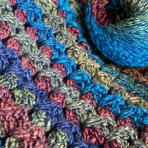 May include: A crocheted blanket featuring a textured cluster stitch pattern in a variegated color palette of deep navy blue, forest green, and rustic burgundy. A matching ball of yarn sits on top of the fabric, displaying the same multi-colored fibers.