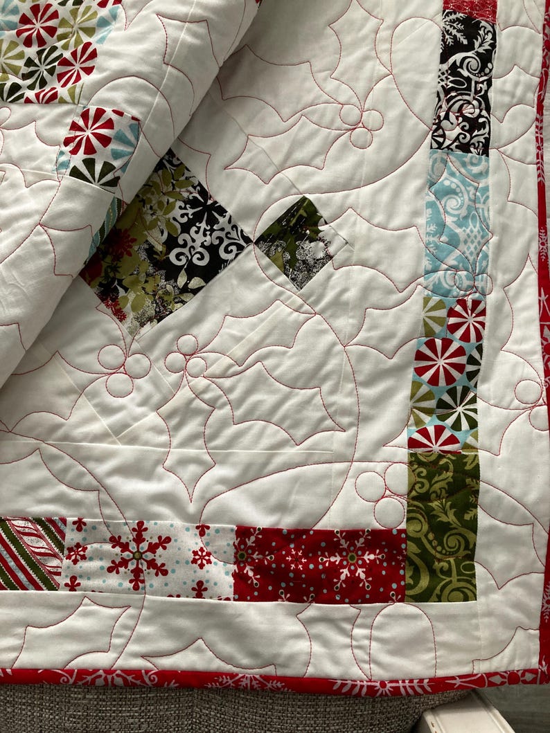 May include: A white quilt with a red and green holly leaf pattern. The quilt is made of various fabrics with different patterns, including snowflakes, candy canes, and geometric designs.
