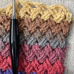 May include: A black crochet hook rests on a colorful, textured crochet project. The project features rows of yellow, brown, gray, pink, and purple yarn, creating a chevron pattern. The hook is made of metal and has a black handle.
