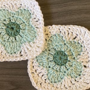 May include: Two square crochet coasters. Each coaster features a floral design in shades of green and white. The coasters are handmade with a textured stitch, perfect for protecting surfaces from hot or cold beverages.
