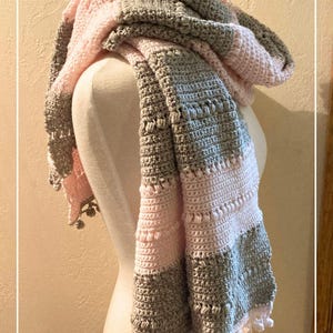 Op de afbeelding: Een grijze en roze gehaakte sjaal met een franje rand. De sjaal is gedrapeerd over een witte mannequin.