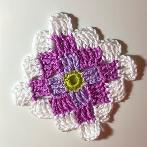 Puede incluir: Una flor tejida a crochet con un borde blanco y un centro hecho de hilo morado y lavanda. El centro de la flor tiene un pequeño círculo verde.