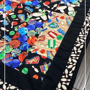 Peut inclure: Un chemin de table matelassé noir sur le thème du bowling. Le chemin de table présente un design en patchwork avec différents motifs de boules et de quilles de bowling. La bordure est un motif de quilles de bowling blanches et rouges.