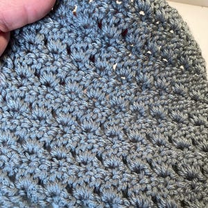 May include: A grey crochet fabric featuring a repeating shell stitch pattern. The textile is crafted from yarn with a subtle sheen, creating a textured surface with small, uniform gaps between the stitches throughout the entire piece.