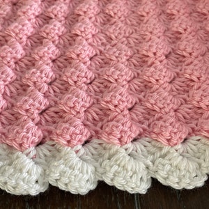 Può includere: Una copertina per neonati in crochet rosa con un bordo in crochet bianco. La copertina ha un motivo testurizzato a forma di diamante.