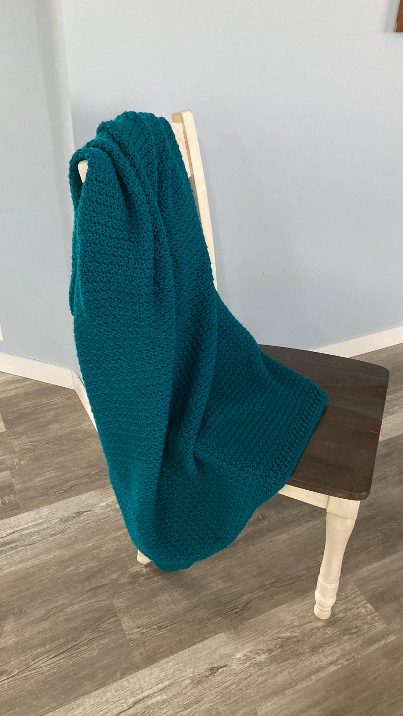May include: A teal blue crocheted throw blanket draped over a wooden chair with white legs.
