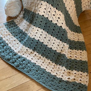 May include: A hand-crocheted blanket with a striped design in teal and white. The blanket is displayed, highlighting its soft texture. A ball of white yarn is placed nearby, indicating its handmade nature. The blanket is approximately 150 cm x 180 cm.