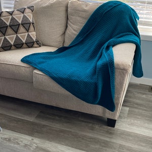 May include: A teal blue throw blanket draped over a beige couch with a patterned pillow. The couch is in a room with wood-look flooring and windows with white blinds.