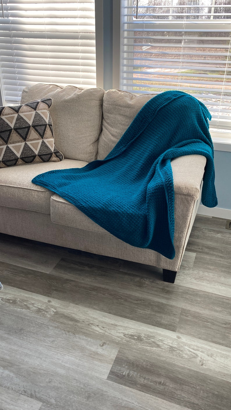 May include: A teal blue throw blanket draped over a beige couch with a patterned pillow. The couch is in a room with wood-look flooring and windows with white blinds.