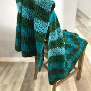 May include: A handmade crochet blanket draped over a wooden chair. The blanket features alternating stripes of teal, turquoise, and forest green. The textured stitches and color combination create a cozy, inviting aesthetic. The blanket is a home decor item.